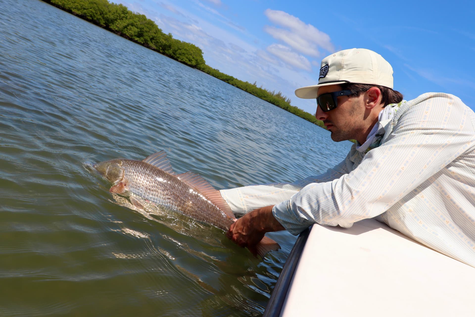 Man wearing hat and sunglasses releasing a fish into calm water from a boat near green shoreline