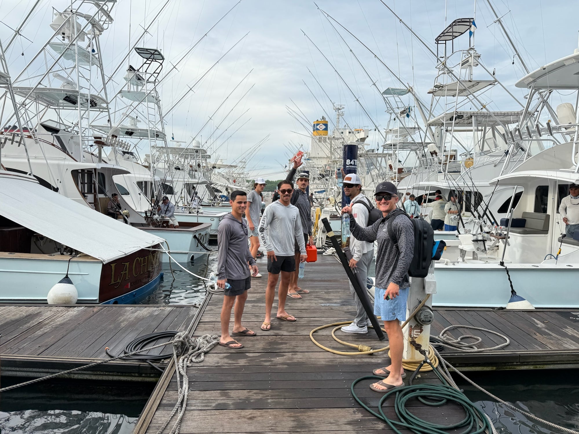 Group of five men standing on wooden dock between sportfishing boats in marina under cloudy sky